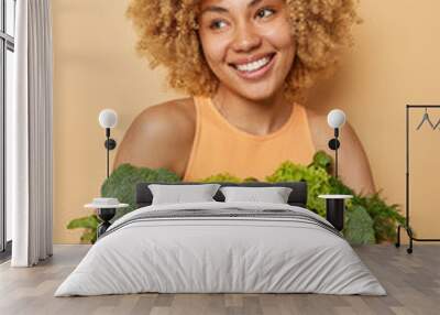 Vertical shot of happy woman poses with fresh green vegetables and fruits keeps to healthy diet wears casual t shirt isolated over brown background. Homegrown grocery. Vegeterian food concept Wall mural