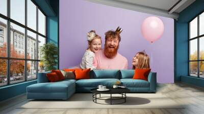 Vertical shot of overemotive joyful single dad and two daughters, celebrate Fathers Day, wear festive outfits, have fun together, isolated over purple background, blank space above for promotion Wall mural