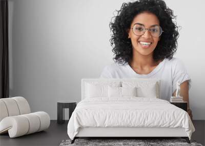 Waist up portrait of beautiful young dark skinned female with positive expression, has Afro hairdo, feels happy after successful bargain, wears big spectacles, isolated over white background Wall mural