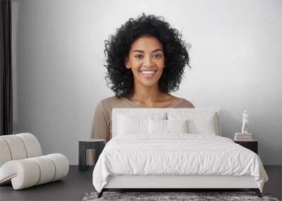 Waist up portrait of cheerful young mixed race female with curly hair posing in studio with happy smile. Dark-skinned woman dressed casually smiling joyfully, showing her white straight teeth Wall mural