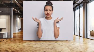 Waist up shot of confused Latin woman raises her hands with palms open conveying sense of hesitation doubt unable to make decision makes difficult choice wears casual t shirt isolated on white wall Wall mural