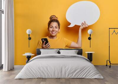 Waist up shot of good looking young European woman with hair bun holds mobile phone and blank communication bubble for your advertising content wears casual t shirt isolated over yellow background. Wall mural