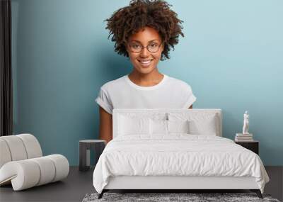 Waist up shot of happy curly woman with toothy smile, wears optical glasses and casual solid white t shirt, expresses good emotions, enjoys nice day, isolated over blue background. Face expressions Wall mural