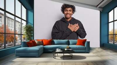 Waist up shot of happy Hindu man with curly hair keeps hands on heart expresses gratitude feels flattered and thankful smiles gladfully wears black sweatshirt isolated over white background. Wall mural