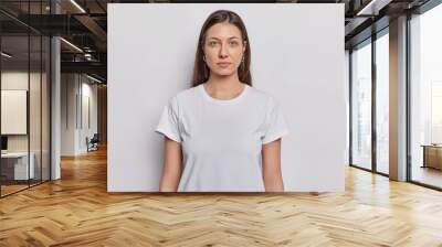 Waist up shot of nice looking woman has dark hair dressed in basic t shirt keeps arms down looks directly at camera isolated on white background. Brunette female model poses in studio for making photo Wall mural