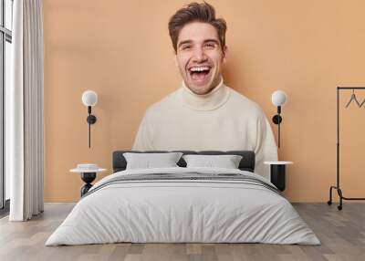 Waist up shot of overjoyed handsome man laughs happily expresses sincere emotions hears something funny dressed in casual white jumper isolated over brown background. People and happiness concept Wall mural