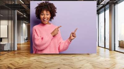 Waist up shot of positive lovely woman with Afro hairstyle, points away with both fore fingers, shows somethig on blank space, has joy expression, wears loose pink jumper, gestures over violet wall Wall mural