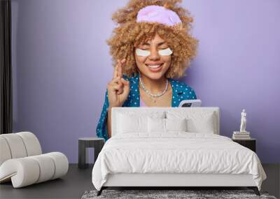 Waist up shot of positive woman keeps fingers crossed makes wish believes in good luck holds mobile phone waits for call wears sleepwear blindfold applies beauty patches under eyes poses indoors Wall mural