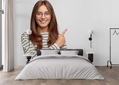 Waist up shot of pretty Caucasian woman with cheerful expression, points with index finger at blank copy space, dressed in striped sweater, shows free space at upper right corner for your promotion Wall mural