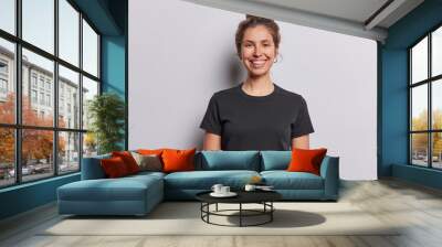 Waist up shot of pretty cheerful European woman with glad smile on face looks directly at camera dressed in casual black t shirt expresses positive emotions isolated over white studio background Wall mural