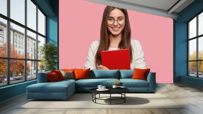 Waist up shot of pretty girl smiles pleasantly, carries red textbook closely, wears white casual jamper, models against pink background. Headshot of Caucasian lady student happy after having classes Wall mural