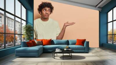 Waist up shot of serious curly haired Hindu man raises palm towards blank space dressed in casual t shirt shows product or advertisement area isolated over brown background. People and promotion Wall mural