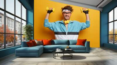 Weight lifting, power and strength concept. Funny active man puts efforts in lifting dumbbells, poses with sport equipment, has workout motivation, wears sport gloves keeps healthy body stays in shape Wall mural