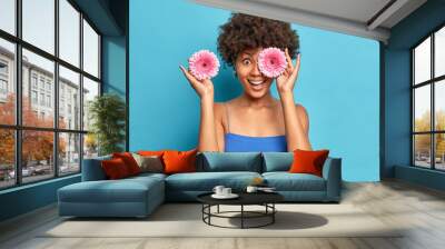 Woman likes fragrant flowers pickes two pink gerberas covers eyes has fun enjoys pleasant aroma dressed in blue dress poses indoor. Afro American florist arranges bouquet for special occasion Wall mural