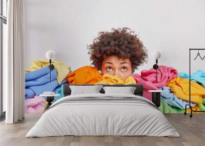 Wondered curly haired ethnic woman focused above surrounded by multicolored laundry cluttered with clothes collects clothing for recycling isolated over white background. Organize your closet Wall mural