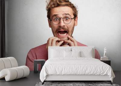 Worried scared fashionable man looks with terrified expression, keeps arms on chin, dressed in pink sweater, isolated over white background. Emotional bearded young guy receives unexpeted news Wall mural