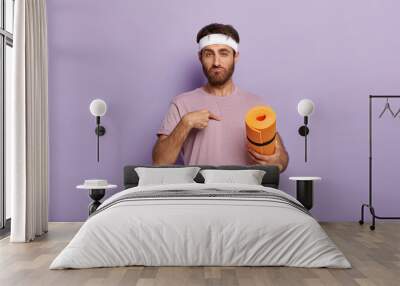 You mean me? Confused unshaven guy points at himself, wears white headband, dressed casually, holds yellow fitness mat, asks whether he should conduct workout training, isolated on purple wall Wall mural