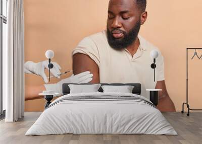 Young bearded ethnic man with dark skin gets injection in arm to stop coronavirus poses against beige background isolated over brown background. Covid 19 vaccine health care and safety concept Wall mural