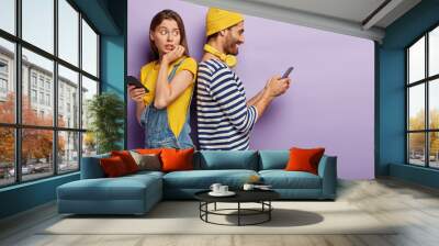 Young couple stand back to back with mobile phones, jealous nervous woman tries to spy what boyfriend is typing, pose against purple background with empty space. Relationship, secrecy, infidelity Wall mural