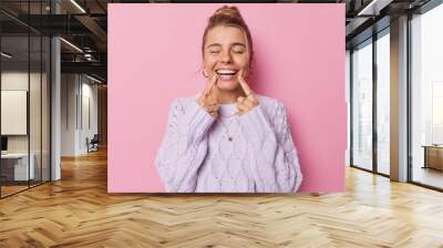 Young glad woman points at toothy smile shows perfect white teeth being in good mood keeps eyes closed dressed in casual knitted jumper isolated over pink background. Positive emotions concept Wall mural