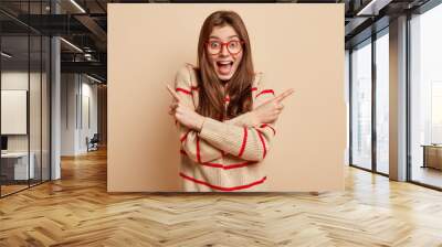 Young happy dark haired woman points sideways, takes decision and makes choice from cool objects, feels uncertain which way to choose, dressed in casual clothing, isolated over beige background Wall mural