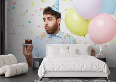 Young man having birthday, receiving congratulations from friends, holding colorful balloons, blowing at candle of cupcake, making wish. Bearded attractive young man coming to birthaday party Wall mural