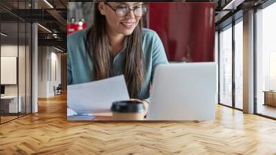 Young woman adminstrative manager reads documentation, has positive expression, looks at laptop computer, drinks takeaway coffee, enjoys cozy atmosphere in kitchen, organizes working process Wall mural