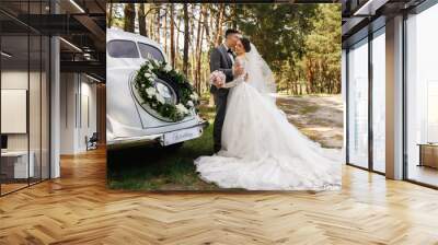 Luxury wedding photography. Elegant wedding couple, groom in grey suit and bride in wedding dress with long sleeves and long train kissing near white Just Married car with inscription 