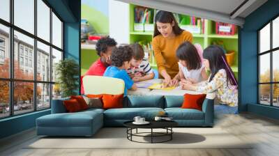 Asian female teacher teaching mixed race kids reading book in classroom,Kindergarten pre school concept. Wall mural