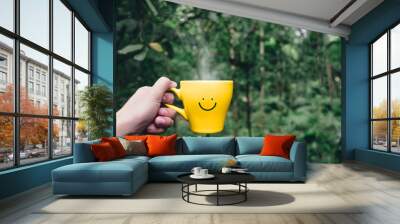 Hand holding hot yellow coffee cup with hand drawn smile face on cup at tropical nature forest,Leisure lifestyle. Wall mural