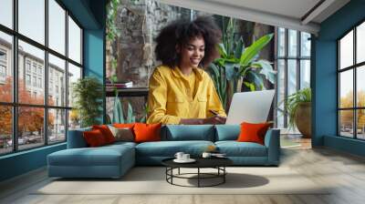 Portrait of young black woman smiling while writing in notebook and using laptop at wooden desk, wearing casual yellow shirt with curly hair sitting near window on the wall with green plant at industr Wall mural