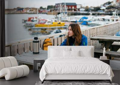 Happy smiling girl traveler drinks warm tea at the rustic wooden table outdoor near the traditional fishing houses Rorbu and fishing ships in Lofoten islands, Norway Wall mural