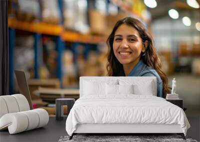 Happy hispanic or latino female young woman warehouse worker sitting at desk with her laptop looking at camera and shelves with cardboard boxes in the logistic distribution delivery centre background Wall mural