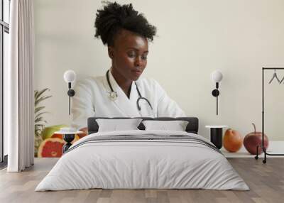 A young African-American nutritionist in a white coat sits at a modern table, writing a recipe, with fresh fruit lying nearby on a white background. Wall mural