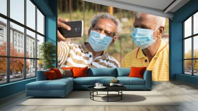 elderly friends with medical face mask taking selfie while sitting at park - concept of happy senior people friendship and meetup after lockdown due to coronavirus covid-19 pandemic. Wall mural