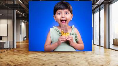 Excited Girl kid with large eyes open, enjoying eating chocolate on blue studio background - concept of unhealthy food consumption and childrens love on chocolate Wall mural