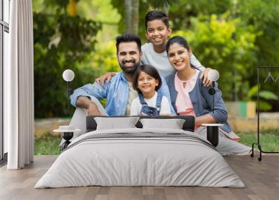 Happy indian couple with kids embracing by looking at camera while sitting at park - concept of relationship, family time and togetherness Wall mural