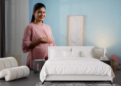 Happy smiling Indian Pregnant woman standing by feeling tummy while looking camera at home - concept of motherhood, expecting baby and parenthood Wall mural