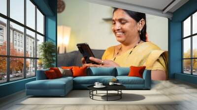 Happy smiling Indian woman busy using mobile phone while sitting on sofa - Concept of people using social media, online messaging app, Internet and entertainment lifestyle. Wall mural