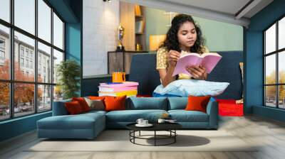 Serious young girl reading book for examination while sitting on sofa at home - concept of education, preparation and hobbies Wall mural