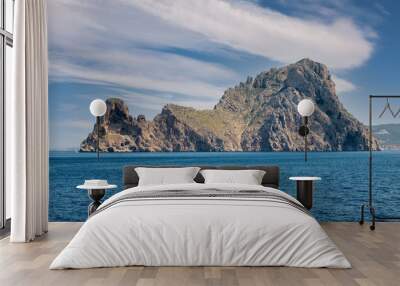 The small island of Vedra off the coast of Ibiza in the Mediterranean Sea. The rock rises steeply from the sea. On the left is a sailing boat. It's a sunny midday in summer with interesting clouds. Wall mural