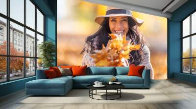 A cheerful brunette in stylish autumn clothes holds an arm full of colorful fallen leaves. Wall mural