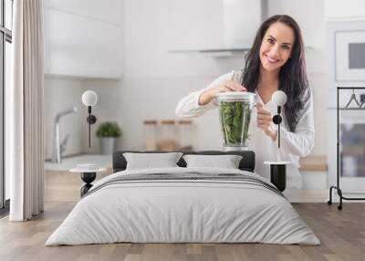 Beautiful lady blends greens in a smoothie mixer with more leaves inside the glass bowl next to her Wall mural