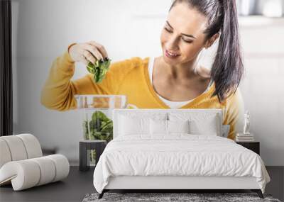 Smiling woman making spinach smoothie, putting leaves in blender Wall mural