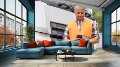 Truck driver in orange vest stands in front of his lorry, testing on a mobile phone Wall mural