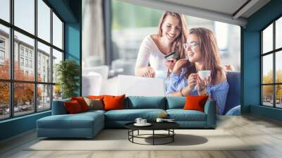 Two young cheerful women in a coffee shop with a credit card and and laptop comfortably buy them over the internet. Wall mural