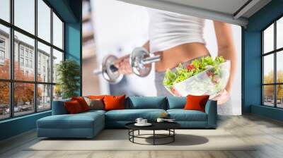 Woman holding bowl from fresh vegetable salad and dumbbell. Wall mural