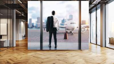 A businessman looking at an airplane through large airport windows, contemplating travel against an urban skyline Wall mural