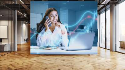 Attractive happy young european businesswoman talking on phone at desk with laptop and growing blue forex chart and index on blurry background. Trade and financial growth concept. Double epxosure. Wall mural