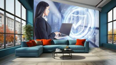 Portrait of attractive young european businesswoman with laptop standing in blurry office interior with glowing shield hologram. Protection and internet firewall concept. Double exposure. Wall mural